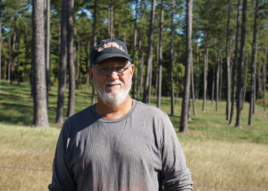 A portait shot of a man in a baseball hat. Behind him are trees and green grass