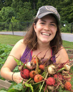 Jess Mrugala contact photo of her smiling holding freshly plucked vegetables from the farm