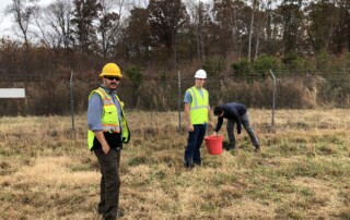 Two men with hard hats and vests looking at the camera. A person is next to them leaning down. They are all in a field of brown grass and behind them is a fence