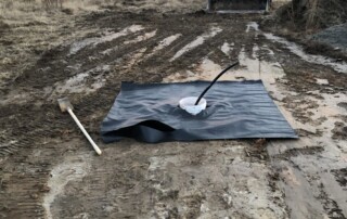 A compact loader in the middle ground coming towards the camera. In the middle ground there is a black tarp covering dirt and a bucket stuck in the middle of the tarp. The land is muddy dirt