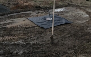 A trailer in the background with trees. In the middle ground there is a black tarp covering dirt and a bucket stuck in the middle of the tarp. A metal pole is in the bucket. The land is muddy dirt