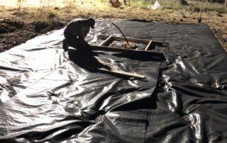 It is nighttime and a person is kneeling in the middle ground. In the middle ground there is a black tarp covering dirt and a bucket stuck in the middle of the tarp. A metal pole is in the bucket