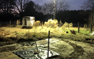 It is nighttime and there is a trailer in the background with trees and a fence. On the near side of the fence is dirt. A black tarp covers a square of the ground. A bucket is in the middle of the tarp and a pole is in the bucket
