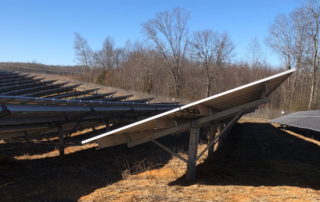 The photo is angled at a solar panel straigh up, the underside of the panel is visible. This panel is in the center of the photo and to the left and right of this panel are other solar panels. In the background are barren trees and blue sky