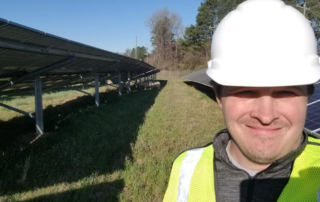 A selfie from a man in a hard hat and yellow vest on the right of the photo. Behind him are two large solar panels and in the background are trees
