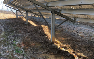 Underside view of a solar panel, looks similar to the underside of metal bleachers. There are metal panels running up and down. The solar panel is in a barren field