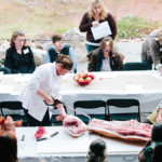 person cutting large piece of meat with meat saw in front of outdoor crowd