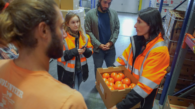 People in orange safety jackets inside food facility. One lady holding a cardboard box full of oranges