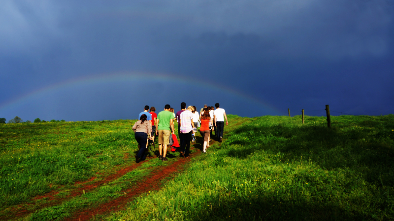 Group of seemingly young adults walking up trail on a grassy hill toward a rainbow in the cool-dark blue sky